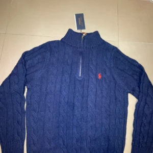 Mörkblå stickad tröja från Ralph Lauren - Säljer en snygg mörkblå stickad tröja från Ralph Lauren med dragkedja och polokrage. Tröjan har ett klassiskt flätmönster och den ikoniska röda loggan på bröstet. Perfekt för kyliga dagar!