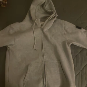 Grå hoodie från Stone Island - Säljer en stilren grå hoodie från Stone Island med dragkedja och justerbar huva. Tröjan har en ikonisk logga på ärmen och praktiska fickor framtill. Perfekt för en avslappnad stil.