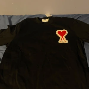 Svart t-shirt med hjärtdetalj från AMI - Snygg svart t-shirt från AMI med en röd hjärtdetalj och bokstaven 'A' på bröstet. Perfekt för en stilren look.