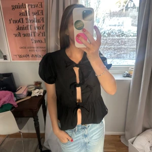 Svart blus med knytband - Trendig svart blus med korta puffärmar och knytband framtill. Passar så bra med alla typer av underdelar✨✨