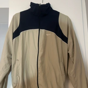 Beige och marinblå vindjacka från Nike - Snygg och sportig vindjacka från Nike i beige och marinblått. Jackan har en dragkedja framtill och en hög krage. Perfekt för en casual look eller träning.