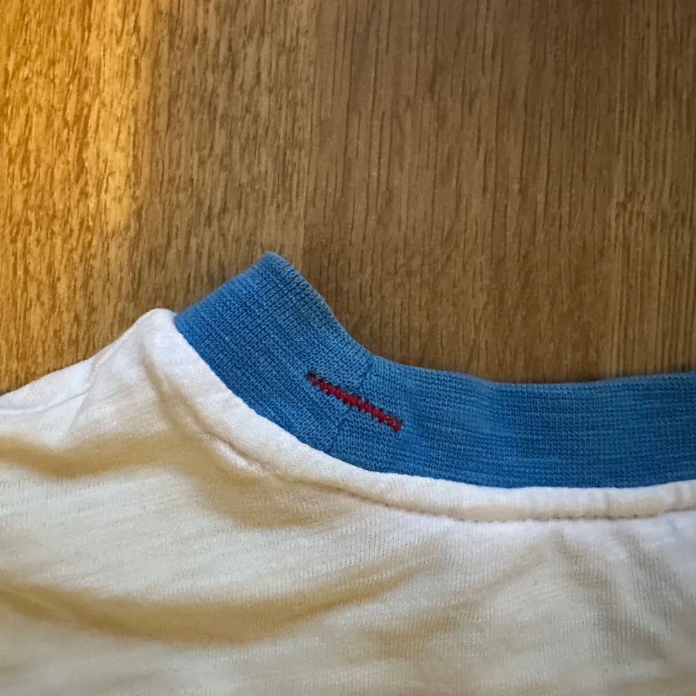 Snygg vit t-shirt från Guess med färgglada ränder och logga på framsidan. Blå detaljer vid krage och ärmslut ger en fräsch look. Lite oversized. Röd tråd för att sy ihop kragen. Ej gjort av mig men sänker priset pga detta.. T-paidat.