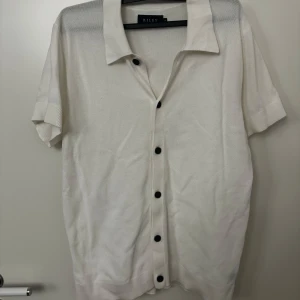 Vit kortärmad skjorta från Riley strl M - Säljer en stilren vit kortärmad skjorta från Riley. Skjortan har svarta knappar och en klassisk krage. Perfekt för en avslappnad men ändå uppklädd look.