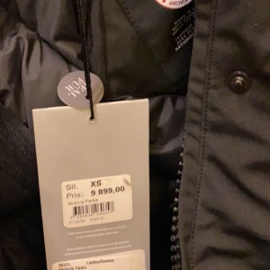 Svart parkajacka med pälsdetalj - Säljer en svart parkajacka från Canada Goose med pälsdetalj på huvan. Jackan har en klassisk design med dragkedja och knappar framtill. Perfekt för kalla vinterdagar.