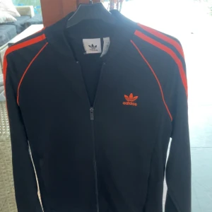 Svart träningsjacka från Adidas - Snygg svart träningsjacka från Adidas med orangea detaljer, säljer då jag vuxit ur den.