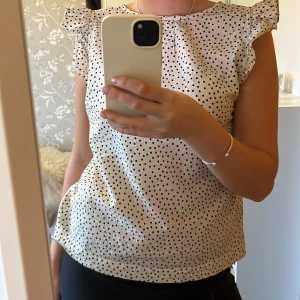 Vit blus med prickigt mönster - Säljer en söt vit blus med små svarta prickar och volangdetaljer vid armarna.