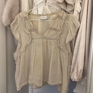 Beige blus från Neo Noir - Charmig beige blus från Neo Noir med volangdetaljer och v-ringning. Blusen har korta ärmar och en luftig passform, perfekt för varmare dagar.