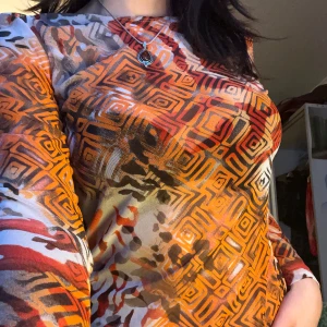 Färgglad mönstrad topp - Säljer en färgglad topp med ett unikt geometriskt mönster i orange, rött och brunt. Toppen har långa ärmar och en figurnära passform. Perfekt för att liva upp garderoben med lite färg och mönster. 