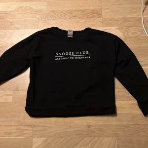 Svart tröja från Kappahl - Säljer en svart tröja från Kappahl med texten 'Snooze Club - Allergic to Mornings' tryckt på framsidan. Tröjan har långa ärmar och en avslappnad passform, perfekt för en chill dag.