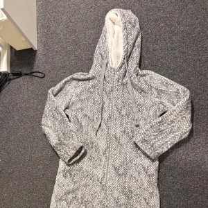 Grå onesie från Hunkemöller - Mysig grå onesie från Hunkemöller med ett fiskbensmönster. Den har en dragkedja framtill och en huva med mjukt foder. Perfekt för kalla dagar hemma.