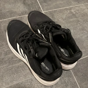 Svarta Adidas sneakers med vita detaljer - Säljer ett par svarta Adidas sneakers med vita ränder och snörning. Skorna har en bekväm sula och är perfekta för vardagsbruk. De har en klassisk design med Adidas-loggan. Skorna har använts varsamt o är i fint skick utan hål i sulorna. Ett litet slitage finns på höger sneakers hälkappa.