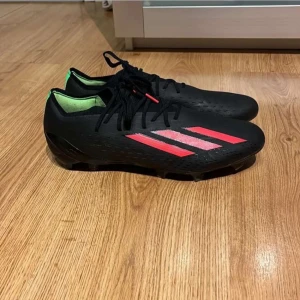 Svarta fotbollsskor med röda detaljer - Snygga svarta fotbollsskor från Adidas med röda ränder och gröna insidor. Skorna har snörning och en slitstark sula perfekt för spel på gräs. Lite slitna