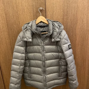 Grå pufferjacka med huva - Snygg grå pufferjacka med avtagbar huva och dragkedja framtill. Jackan har en logotyp på ärmen och justerbara snören vid huvan. Väldigt varm och bra för kalla väder. Jackan kan justeras till din passform vid midjan huvan och handlederna. 