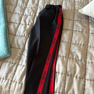 Svarta byxor med röd Voltaire-detalj - Snygga svarta kostym byxor med en röd sidodetalj där det står 'Voltaire'. Byxorna har en elastisk midja för extra komfort och en modern look. Perfekta för en avslappnad stil med en touch av färg. Från Zadig & Voltaire