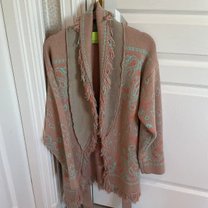 Alanui inspirerad kofta från Paolo Petrone  - Säljer en mysig beige kofta med ett färgglatt mönster i turkos och rosa. Koftan har långa ärmar och fransar längs kanterna samt ett knytband i midjan. Perfekt för en bohemisk stil.