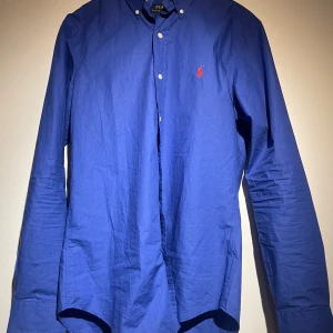 Blå skjorta från Ralph Lauren - Snygg blå skjorta från Ralph Lauren med röd broderad logga på bröstet. Skjortan har en slim passform och är långärmad med knappar framtill. Perfekt för en stilren look.