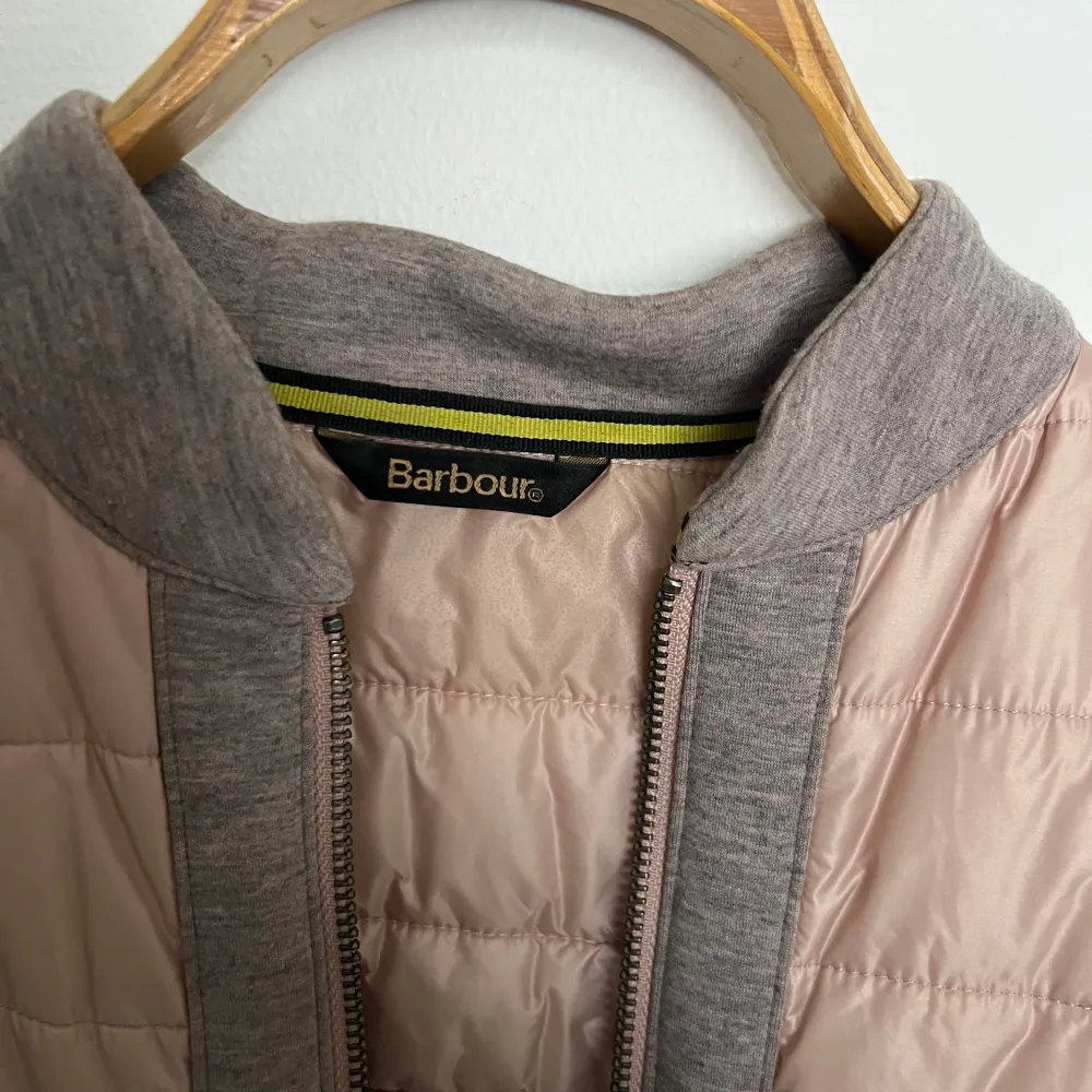 Snygg jacka från Barbour i en kombination av grått och rosa. Jackan har en quiltad framsida och rygg i rosa, med gråa ärmar och krage. Den är försedd med dragkedja och har en modern look som passar perfekt för vår. . Takit.