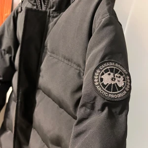 Jacka Canada Goose Macmillan Parka Black label - Säljer en svart dunjacka från Canada Goose med en broderad logotyp på ärmen. Jackan har dragkedja och knappar framtill samt en justerbar huva. Perfekt för kalla vinterdagar. Passar För S och M