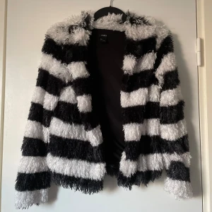 Randig pälsjacka från Lindex - Säljer en supermjuk pälsjacka från Lindex i svart och vitt. Jackan har ett randigt mönster och är långärmad, perfekt för att sticka ut i mängden. Den är gjord i ett syntetmaterial som ger en mysig känsla.
