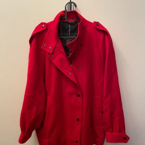 Röd ullkappa från Burberry - Säljer en snygg röd kappa från Burberry i 100% ull med ett klassiskt rutigt foder i bomull. Den har en stilren design med knappar och axelklaffar. Perfekt för höst och vinter!