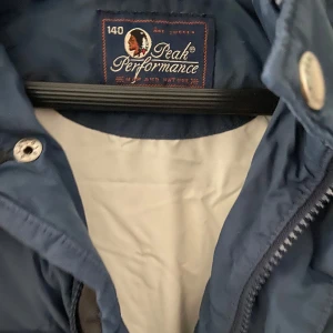Blå dunväst från Peak Performance - Säljer en snygg blå barndunväst från Peak Performance. Västen har en hög krage och stängs med både dragkedja och knappar. Den har två praktiska fickor med dragkedja framtill. Perfekt för kyliga dagar när du vill hålla värmen utan att bära en jacka.