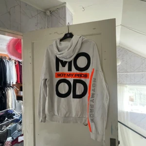 Vit hoodie med tryck - Säljer en vit hoodie med texten 'MOOD' och 'NOT MY PROB' i svart och orange. Den har en stor ficka fram och en bekväm huva. Perfekt för en avslappnad stil!