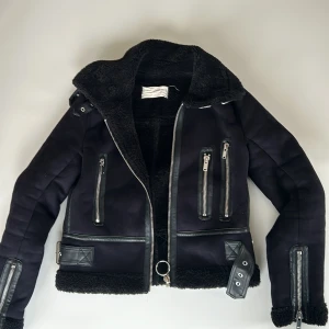 Svart jacka med dragkedjor och bälten - Säljer en snygg svart jacka med coola detaljer som dragkedjor och bälten. Jackan har en mysig foder och är perfekt för kyligare dagar. Den har en stilren design med långa ärmar och en skön passform.