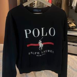 Säljer en snygg svart tröja från Ralph Lauren. Den har en klassisk design med 'POLO' och 'Ralph Lauren' tryckt i vitt och rött på framsidan. Perfekt för en stilren och avslappnad look. Tröjan är långärmad och gjord i ett mjukt material som känns skönt mot huden.