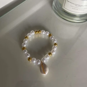 Pärlarmband med snäckdetalj - Snyggt armband med vita pärlor och guldfärgade detaljer. Armbandet har en cool snäckdetalj som hänger ner, vilket ger en somrig och strandig känsla. Perfekt för att piffa upp vilken outfit som helst!