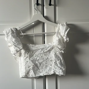 Vit topp från Amisu i storlek S - Säljer en söt vit topp från Amisu med volangdetaljer och broderade mönster. Toppen är croppad och har en elastisk rygg för bättre passform. Passar jättebra med en kjol men även ett par snygga jeans. Perfekt för sommarens varma dagar!