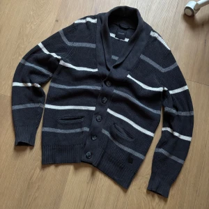 Mörkgrå randig kofta från Blueprint - Säljer en snygg mörkgrå kofta från Blueprint med vita och ljusgrå ränder. Koftan har en sjalkrage och knappar framtill. Perfekt för kyliga dagar! Om du undrar hur den sitter på så kolla min annons med den blåa dobber cardiganen, den sitter samma som den✌️