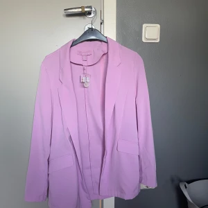 Ljuslila kavaj  - Snygg ljuslila kavaj med klassisk design. Perfekt för att ge en färgklick till din outfit. Kavajen har långa ärmar och två fickor framtill.