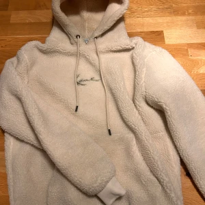 Beige fleece hoodie ll Karl Kani - Mysig beige hoodie i fleece från Karl Kani med broderad logga på bröstet. Den har en stor ficka framtill och justerbar huva med dragsko. Perfekt för kyliga dagar!