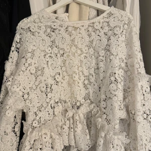 Vit spetsklänning - Elegant vit spetstopp med vackra detaljer och volanger. Toppen har långärmade ärmar en design som passar perfekt för speciella tillfällen.