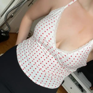 Vit linne med röda hjärtan - Söt vit linne med röda hjärtan och spetsdetaljer längs kanten. Perfekt för en romantisk look. Axelbandslös design som ger en bekväm och luftig känsla.