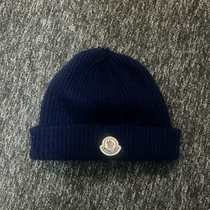 Mörkblå ribbad mössa från Moncler - Snygg mörkblå mössa från Moncler med ribbad design och uppvikt kant. Perfekt för kyliga dagar och ger en stilren look. Moncler-logga framtill.
