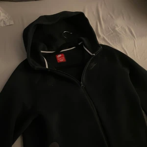 Svart hoodie från Nike/ tech fleece - Säljer en svart hoodie från Nike med dragkedja och huva. Perfekt för en casual look. Den har en diskret Nike-logga på bröstet och vita detaljer vid dragkedjan. Skön passform i stort sätt nyskick.
