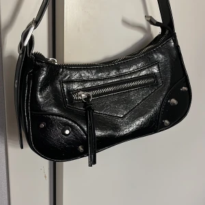 Svart axelväska med nitar - Snygg svart axelväska med nitar och dragkedja framtill. Väskan har en justerbar axelrem med metallhål. Fick den av min kusin så vet inte vart den är ifrån.💕