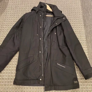 Svart vinterjacka från Peak Performance - Säljer en stilren svart vinterjacka från Peak Performance med huva och dragkedja. Jackan har knappar och praktiska fickor framtill. Perfekt för kyligare dagar. Begagnat skick men hel. Modell Melfort 