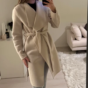 Beige ullkappa med knytband - Snygg beige kappa med knytband i midjan och långa ärmar. Kappan är av ull och i storlek 34. Använd fåtal gånger och i väldigt bra skick. Säljer för jag köpt en ny ullkappa🫶🏼🩷pris kan diskuteras vid snabb affär.   ord.pris 2000kr 