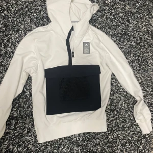 Vit hoodie från Jordan x Paris Saint-Germain - Snygg vit hoodie från Jordan i samarbete med Paris Saint-Germain. Den har en stor svart ficka framtill med dragkedja och ett coolt tryck på ryggen med Jordan-loggan och texten 'Paris'. Perfekt för sportiga tillfällen eller en avslappnad stil.
