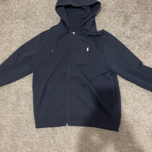 Mörkblå hoodie från Polo Ralph Lauren - Säljer en snygg mörkblå hoodie från Polo Ralph Lauren med dragkedja och huva. Den har en broderad logotyp på bröstet och praktiska fickor framtill. Perfekt för en avslappnad stil.