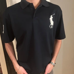 Marinblå pikétröja från Ralph Lauren - Snygg marinblå pikétröja från Ralph Lauren med klassisk krage och korta ärmar. Tröjan har en stor broderad logga på bröstet och siffran 3 på ärmen. Perfekt för en stilren look. Storlek L 