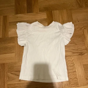 Vit topp från Gina Tricot - Säljer en söt vit topp från Gina Tricot med volangdetaljer på ärmarna. Perfekt för en stilren look. Toppen är i en mjuk och bekväm kvalitet.