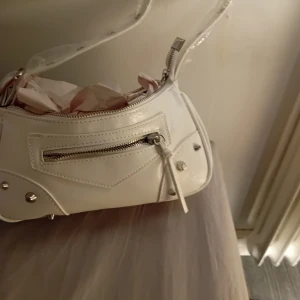 Vit axelväska med dragkedja - Snygg vit axelväska i syntetmaterial med dragkedja och nitar. Perfekt för att ge en stilren touch till din outfit. Väskan har en justerbar axelrem och en praktisk ficka framtill.