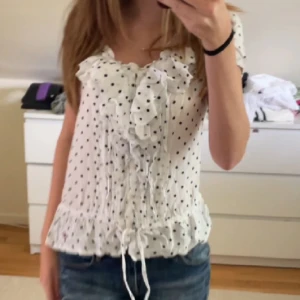 Vit prickig blus med volangdetaljer - Söt vit blus med svarta prickar. Blusen har korta ärmar och en knytning i midjan för en feminin look. Säljer för använder inte. Använd ca 2 gånger.