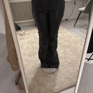 Svarta skinnbyxor - Snygga svarta skinnbyxor med låg midja och bootcut-stil. Perfekta för en trendig look. Byxorna har en slät yta och stängs med dragkedja och knapp framtill.