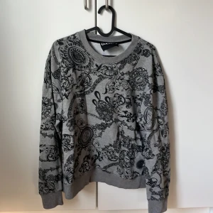 Grå mönstrad tröja från Versace Jeans Couture - Snygg grå tröja från Versace Jeans Couture med ett detaljerat svart mönster. Tröjan har långa ärmar och ribbade muddar vid ärmslut och nederkant. Perfekt för en stilren look. Priset går att diskutera 