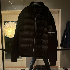 Svart pufferjacka från Bvaldi - Snygg svart pufferjacka från Bvaldi med dragkedja och långärmad design. Jackan har en stilren look med märkets logga på ärmen. Perfekt för kyligare dagar.