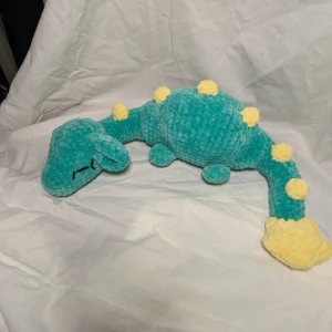 Turkos och gul virkad Nessie✨ - Charmig virkad Nessie i turkos med gula prickar och svans. Perfekt som en mysig leksak eller dekorativ kudde. Den är mjuk och gosig, vilket gör den idealisk för barn eller nessieälskare. Den är egenvirkad i 100% polyester. Längd: 57 cm, höjd: 15 cm, bred: 18 cm